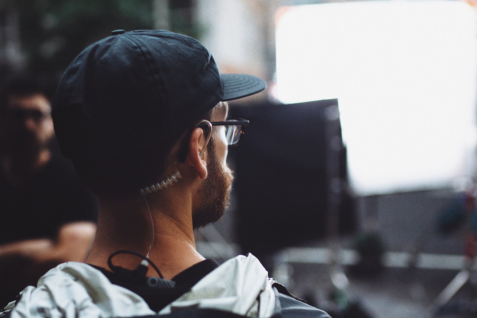 crew member in a cap and glasses seen from behind, earpiece in, monitor ahead