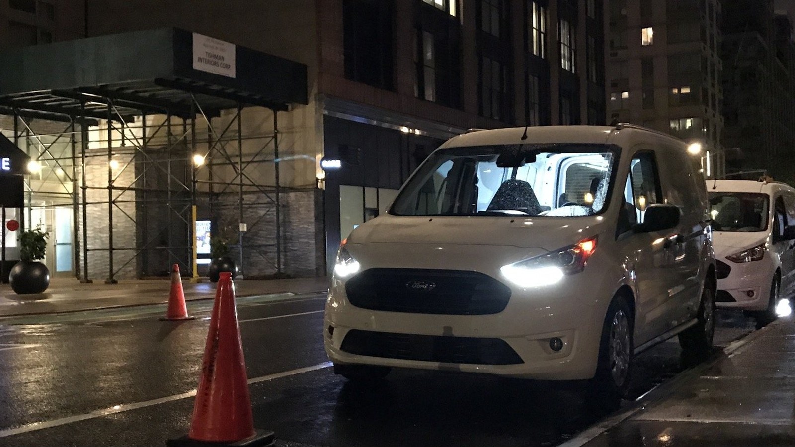 white Ford Transit Connect parked on a wet New York street at night, headlights on, traffic cone in front